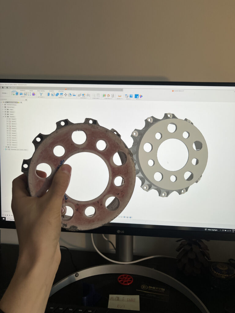 BMW M1 piduriketta originaal adapter/spacer käes, taustal ekraanil 3D-skaneeringu põhjal loodud CAD-mudel.
